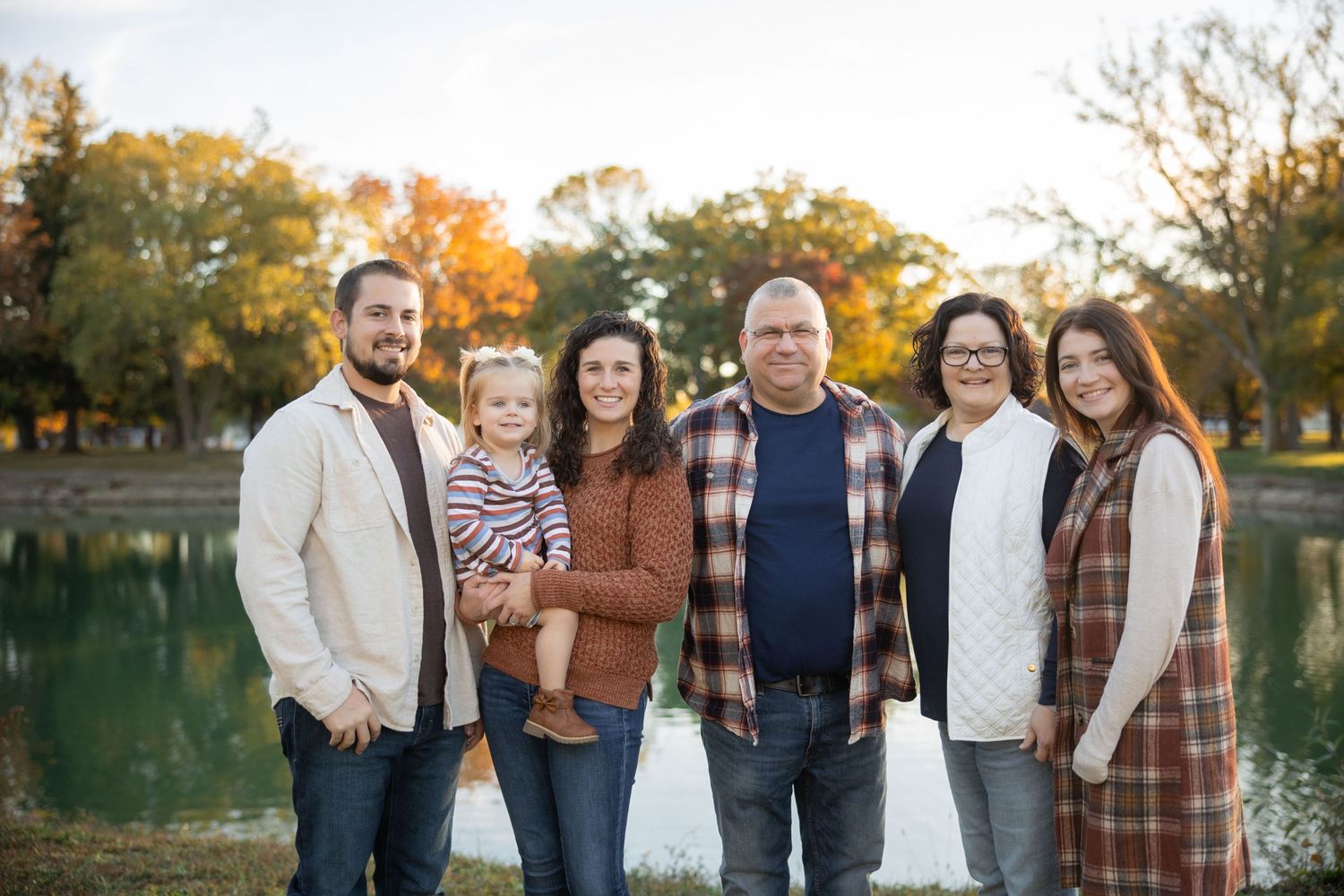 Large family group poses together outdoors near lake during autumn photo session.