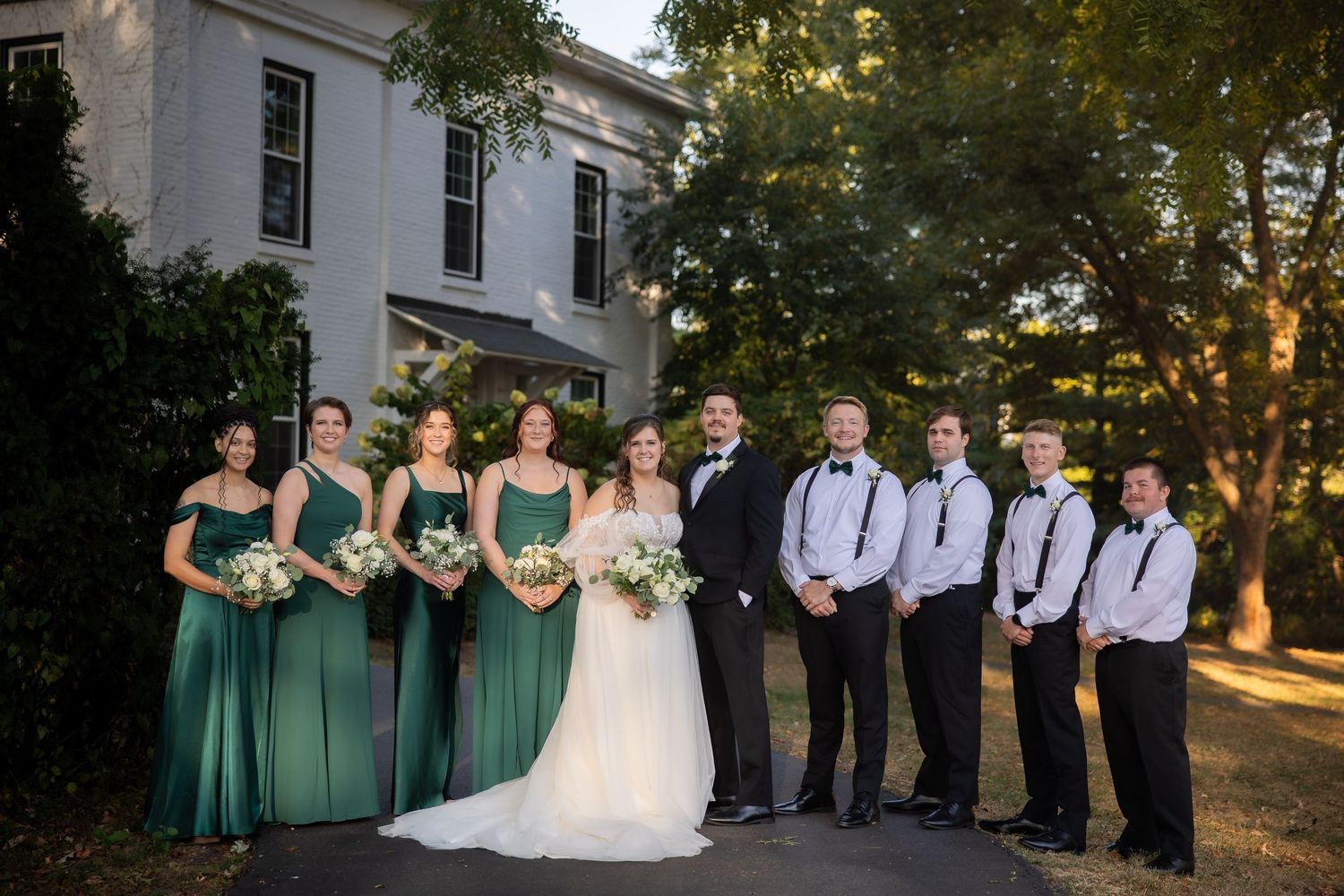 Wedding party in green bridesmaid dresses and black tuxedos posing outside historic venue.
