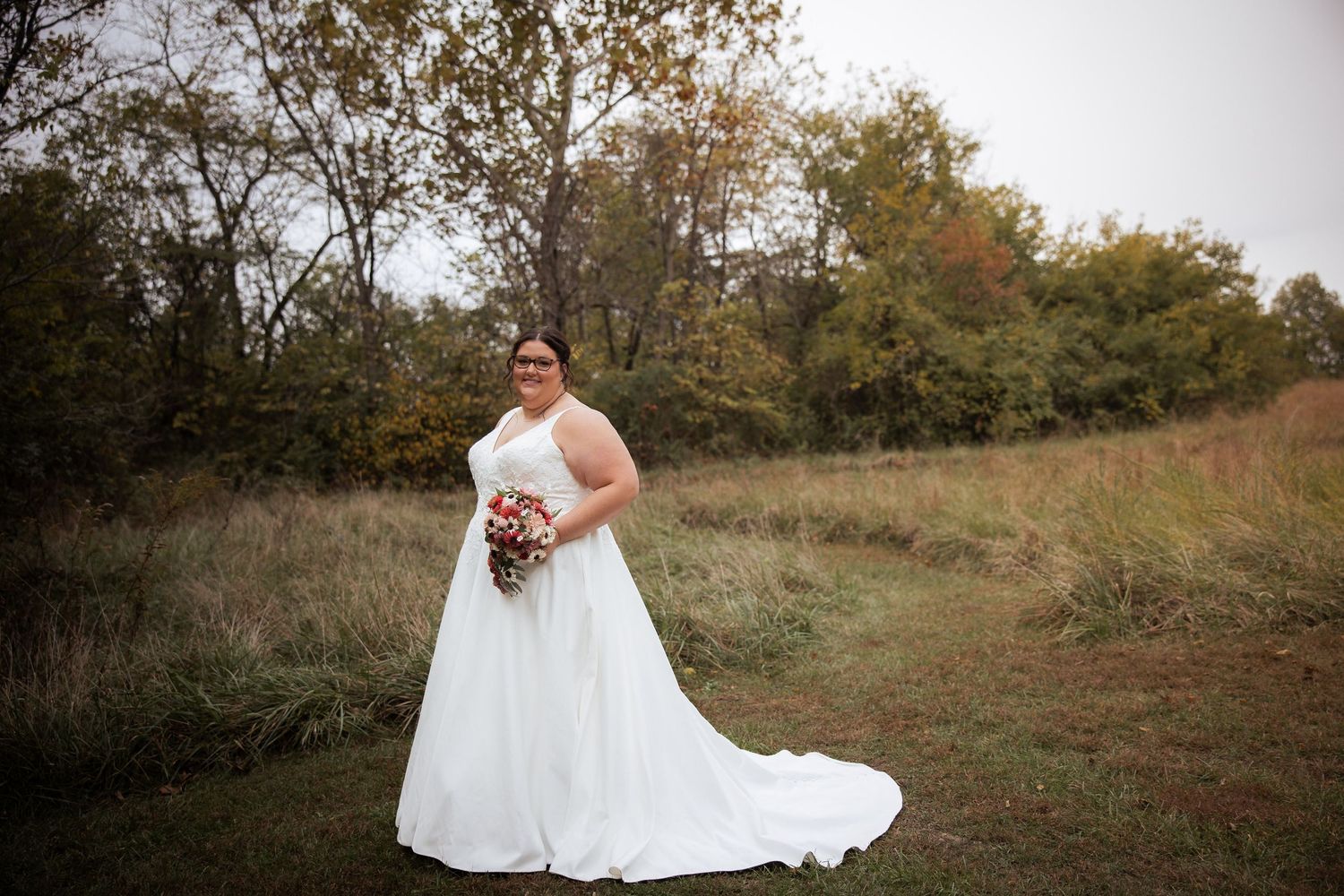 A bride in a flowing white dress poses against a natural hillside backdrop during fall season.
