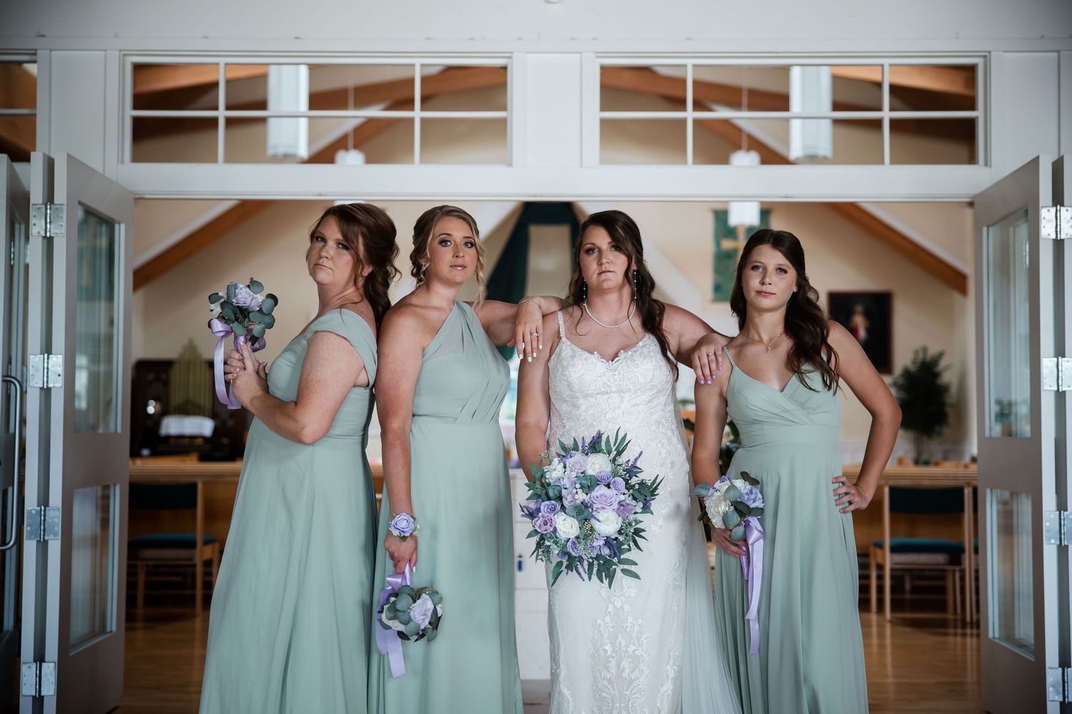 Bride in white lace gown poses with three bridesmaids in sage green dresses holding purple floral bouquets indoors.