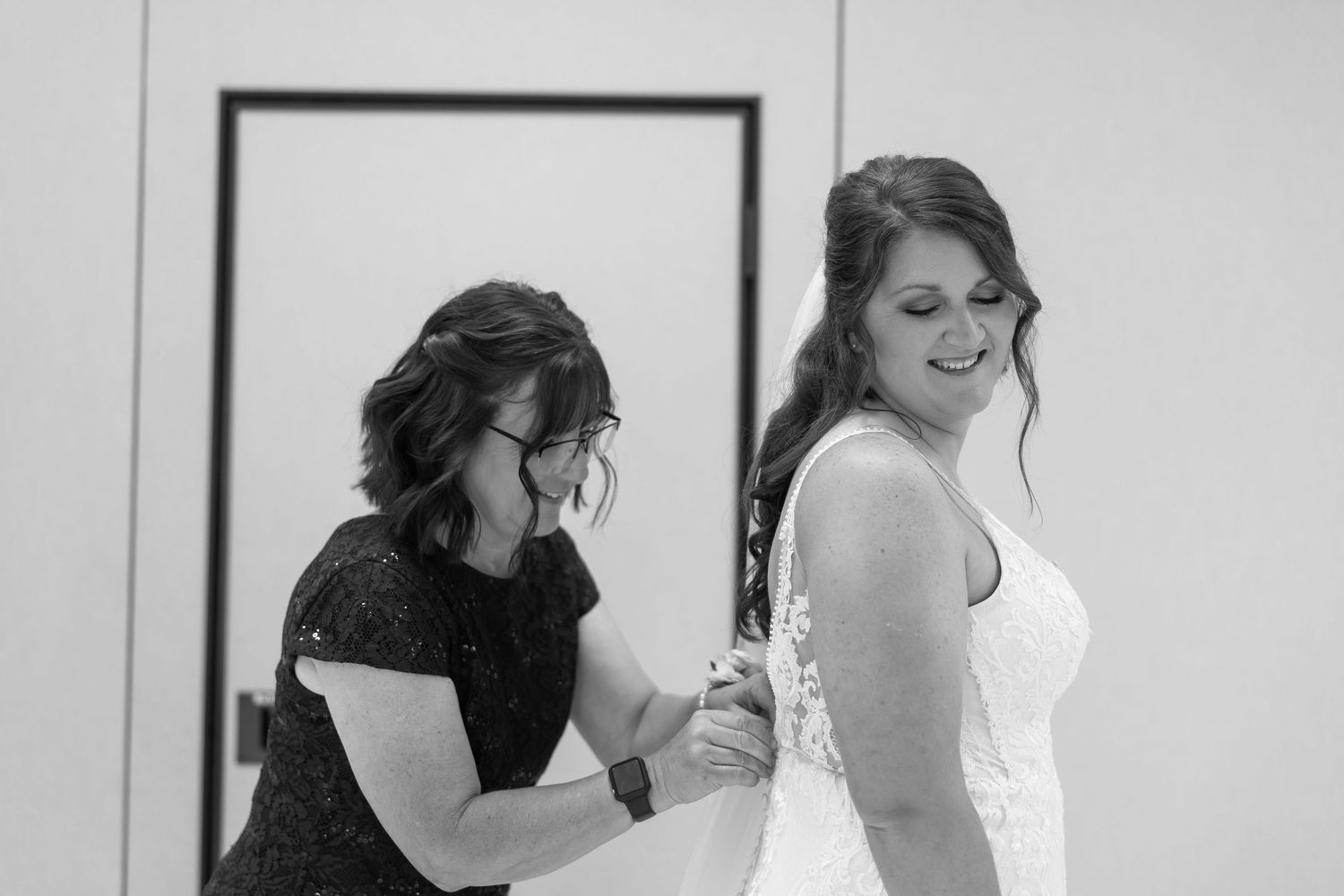 Black and white photo of a woman helping a smiling bride fasten her wedding dress before the ceremony.