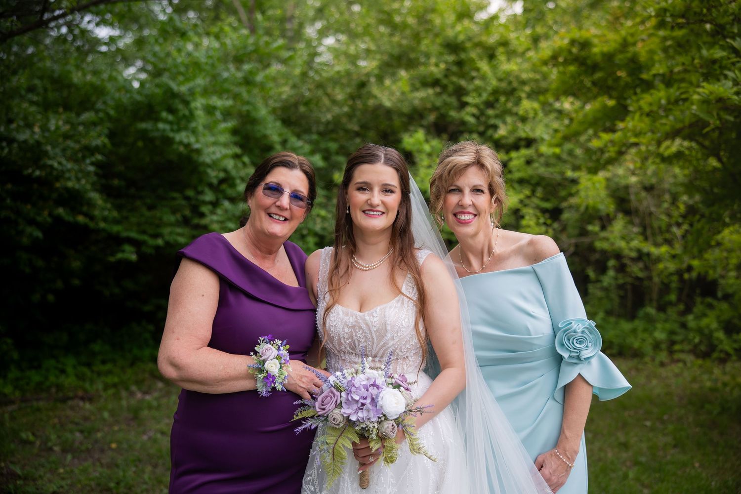 Bride in white lace gown poses with two women in purple and blue dresses holding floral bouquets outdoors.