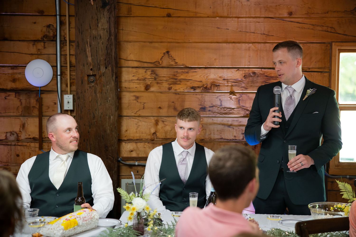 Best man delivers wedding speech holding microphone and champagne glass while groom and groomsman sit at head table.