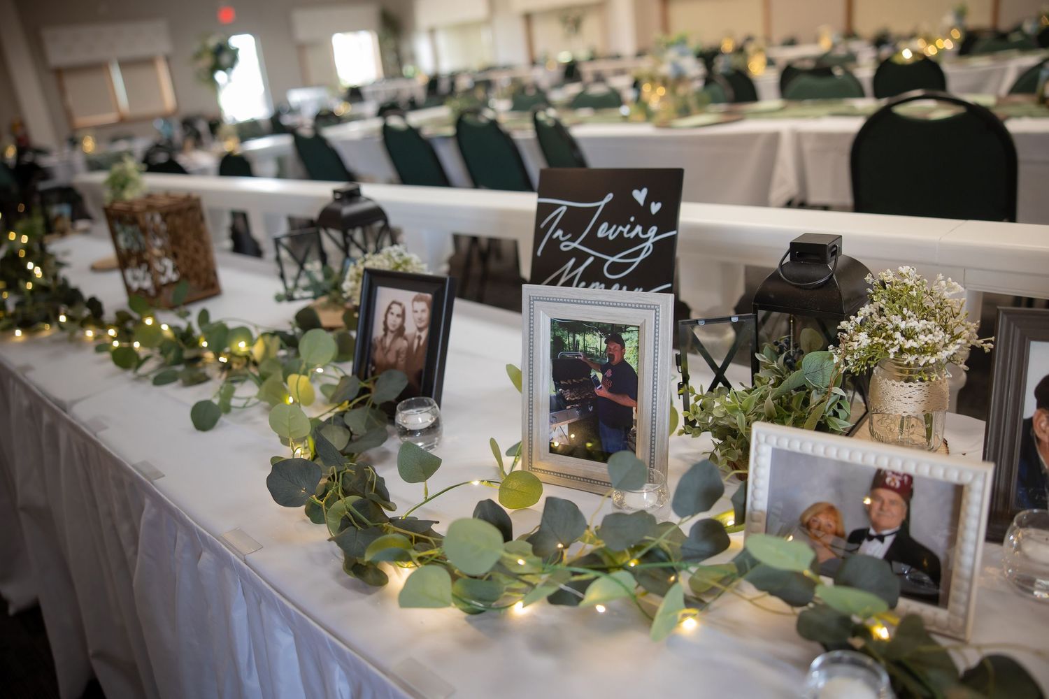 Wedding reception table decorated with eucalyptus garland, fairy lights, framed photos, and lanterns in an elegant banquet hall.