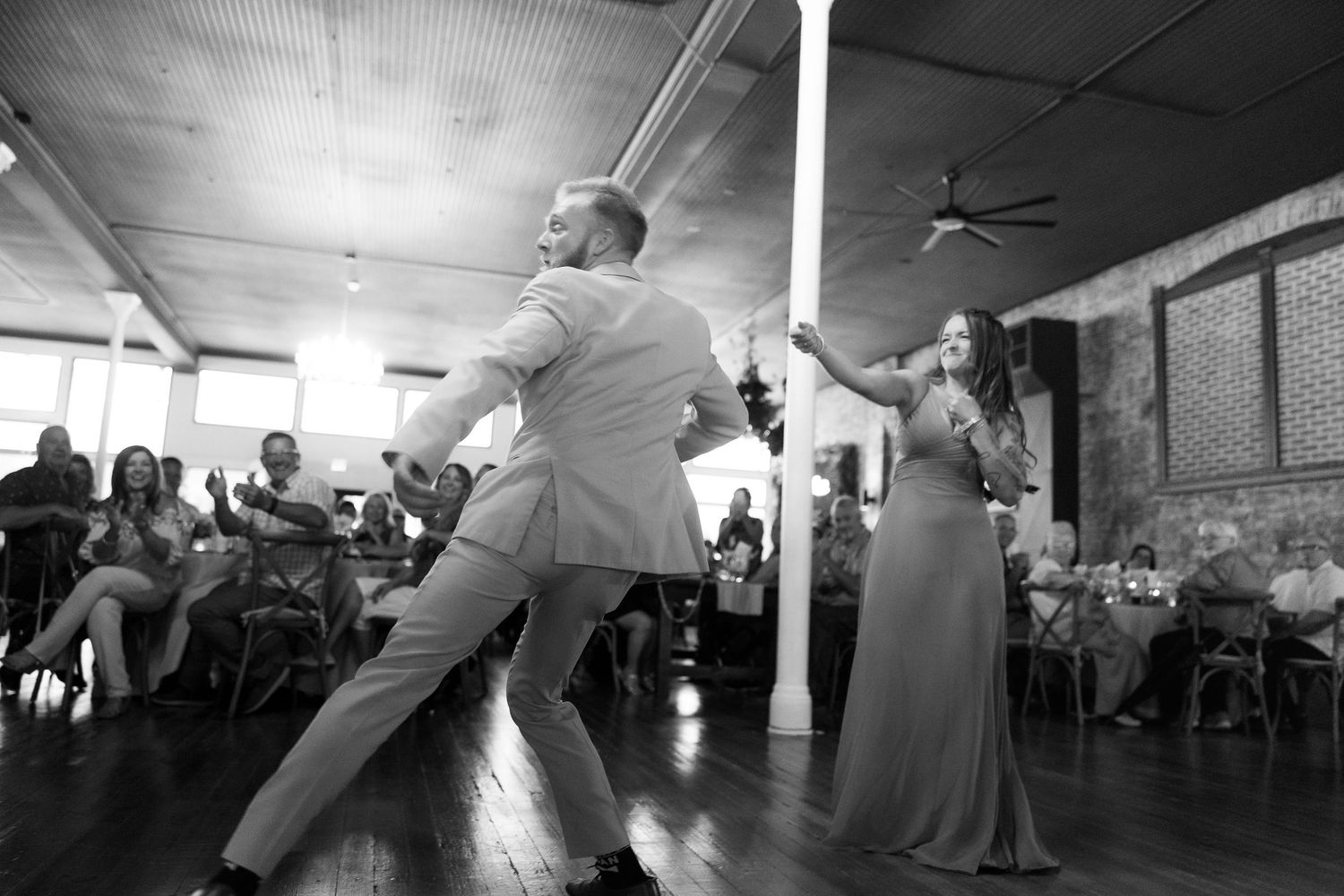 A couple performs an energetic dance move at their wedding reception as guests watch from their tables.