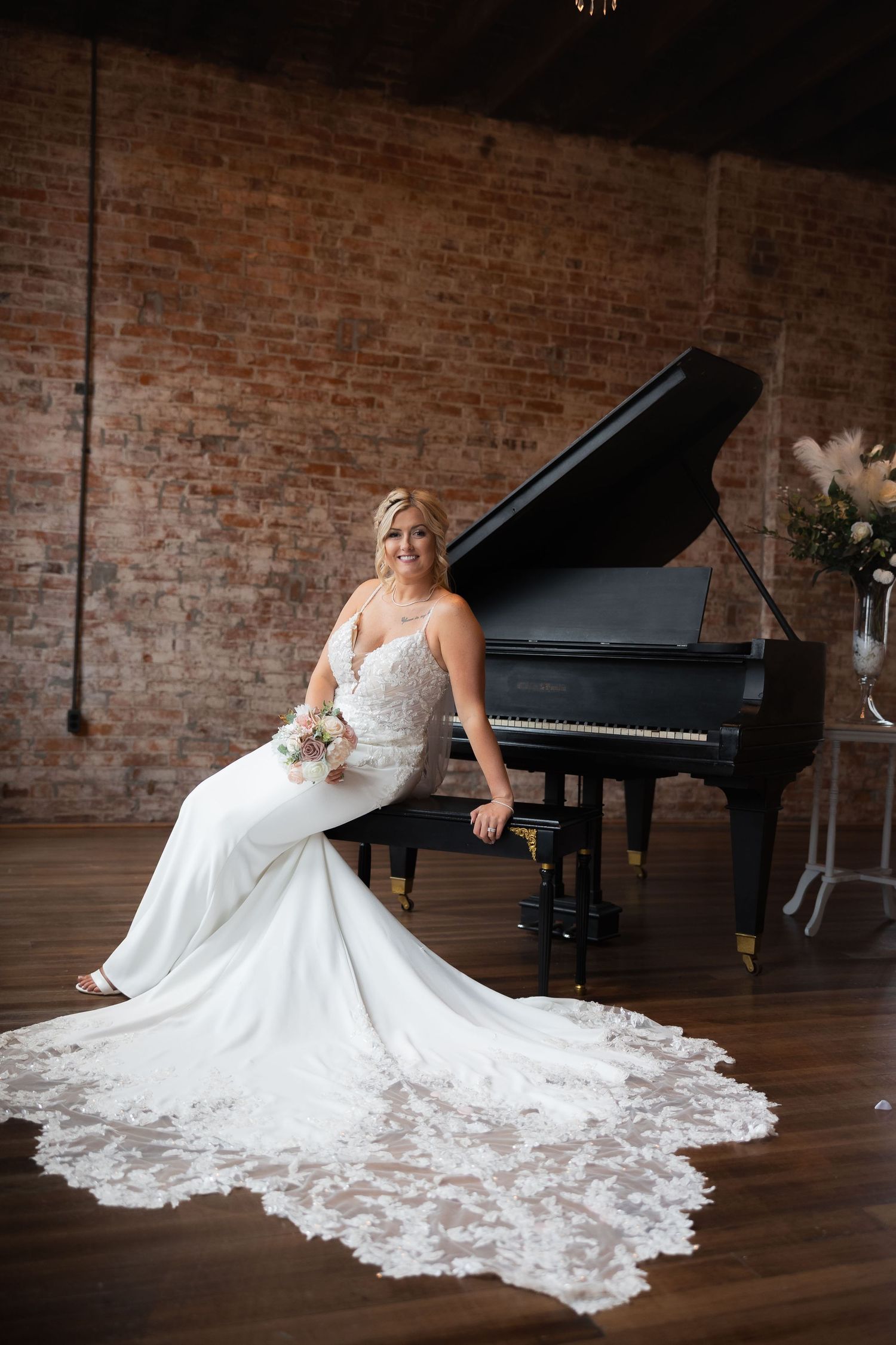 A stunning wedding dress with a long lace train elegantly draped around a black grand piano in a rustic brick venue.