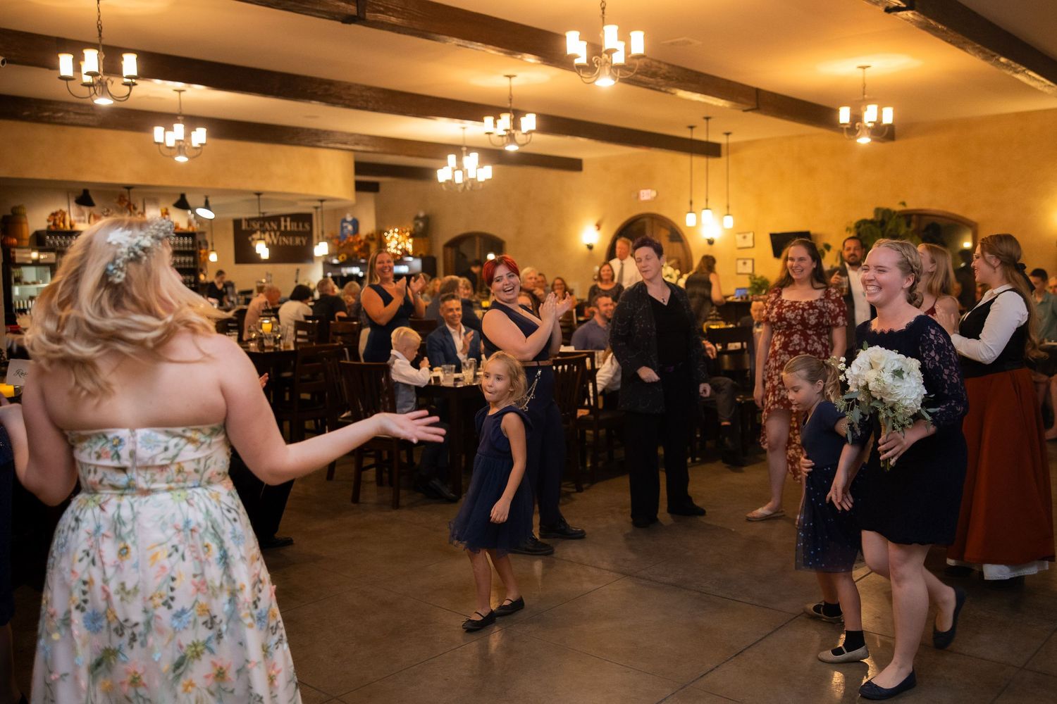 Wedding guests dancing and celebrating in a warmly lit reception hall with exposed beam ceilings.