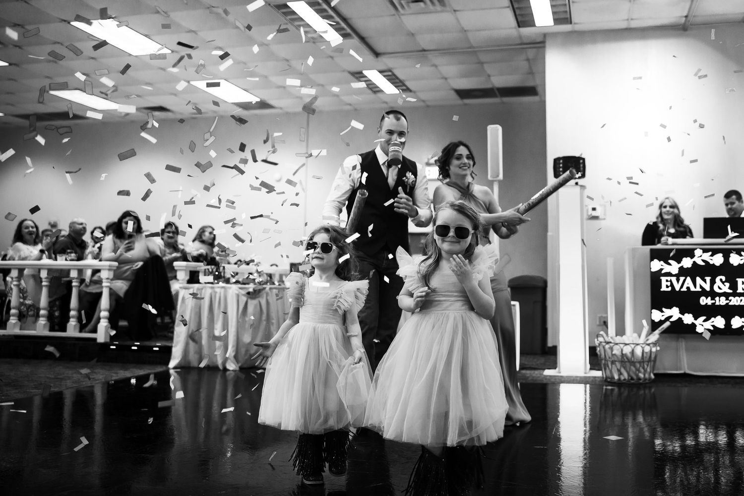 Bride and groom enter wedding reception with flower girls as confetti falls in black and white photo.