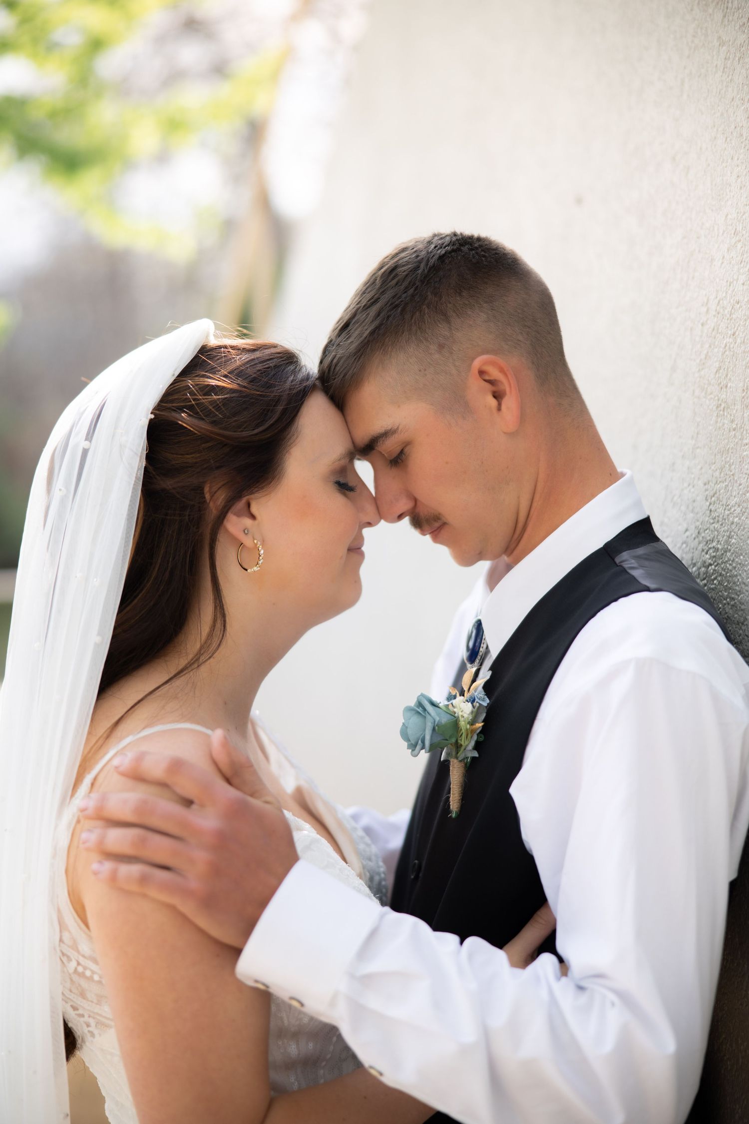 Romantic bride and groom touching foreheads tenderly against white wall at wedding.