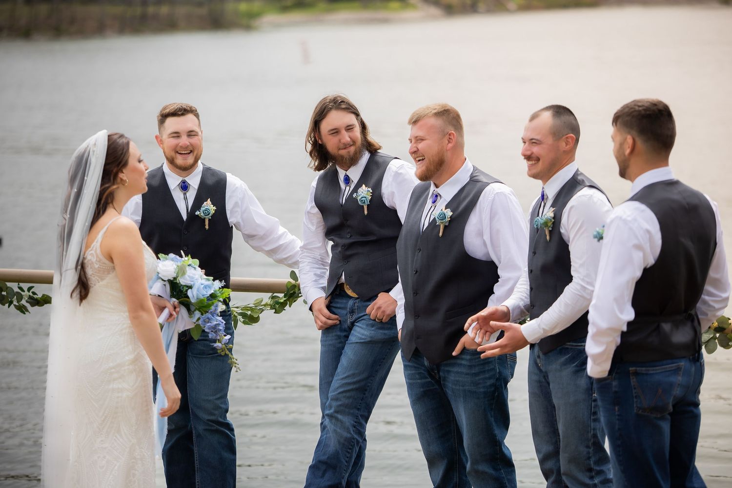 A bride with a bouquet laughs with groomsmen in vests and jeans by a scenic waterfront.