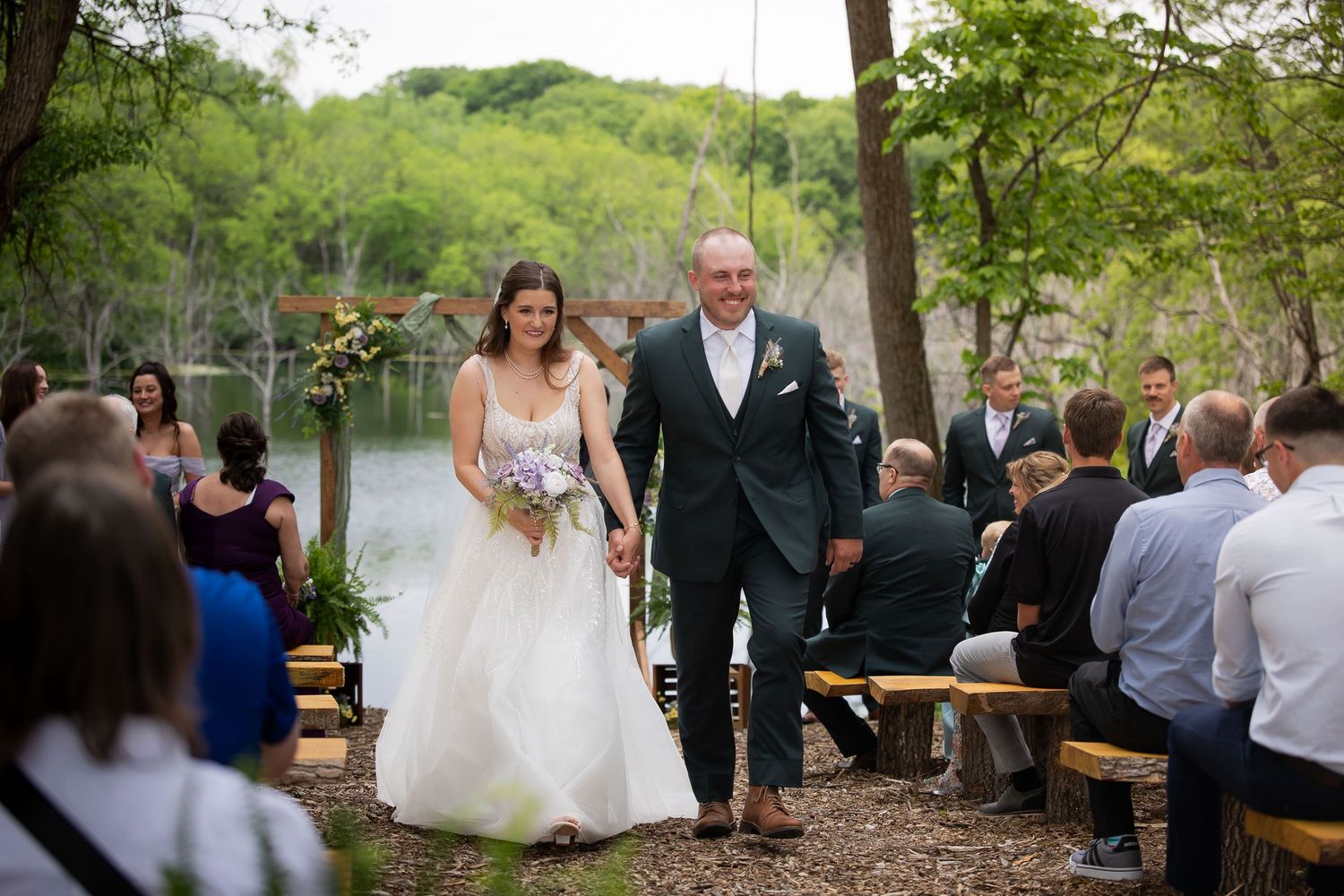 Newlywed couple walks back down aisle hand in hand after outdoor lakeside woodland wedding ceremony.
