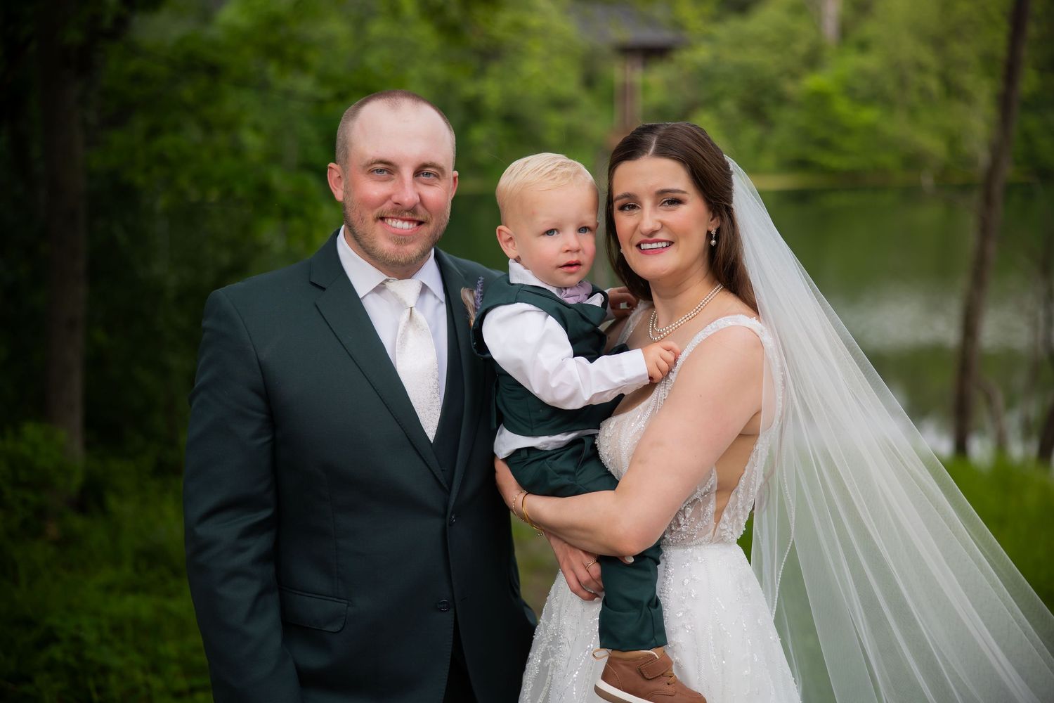 Smiling bride and groom in green suit pose with their toddler son at outdoor lakeside wedding.