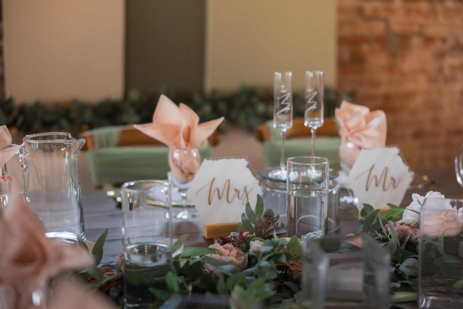 Elegant wedding head table with Mrs. and Mr. signs, blush pink napkins, champagne flutes, and lush greenery centerpieces.