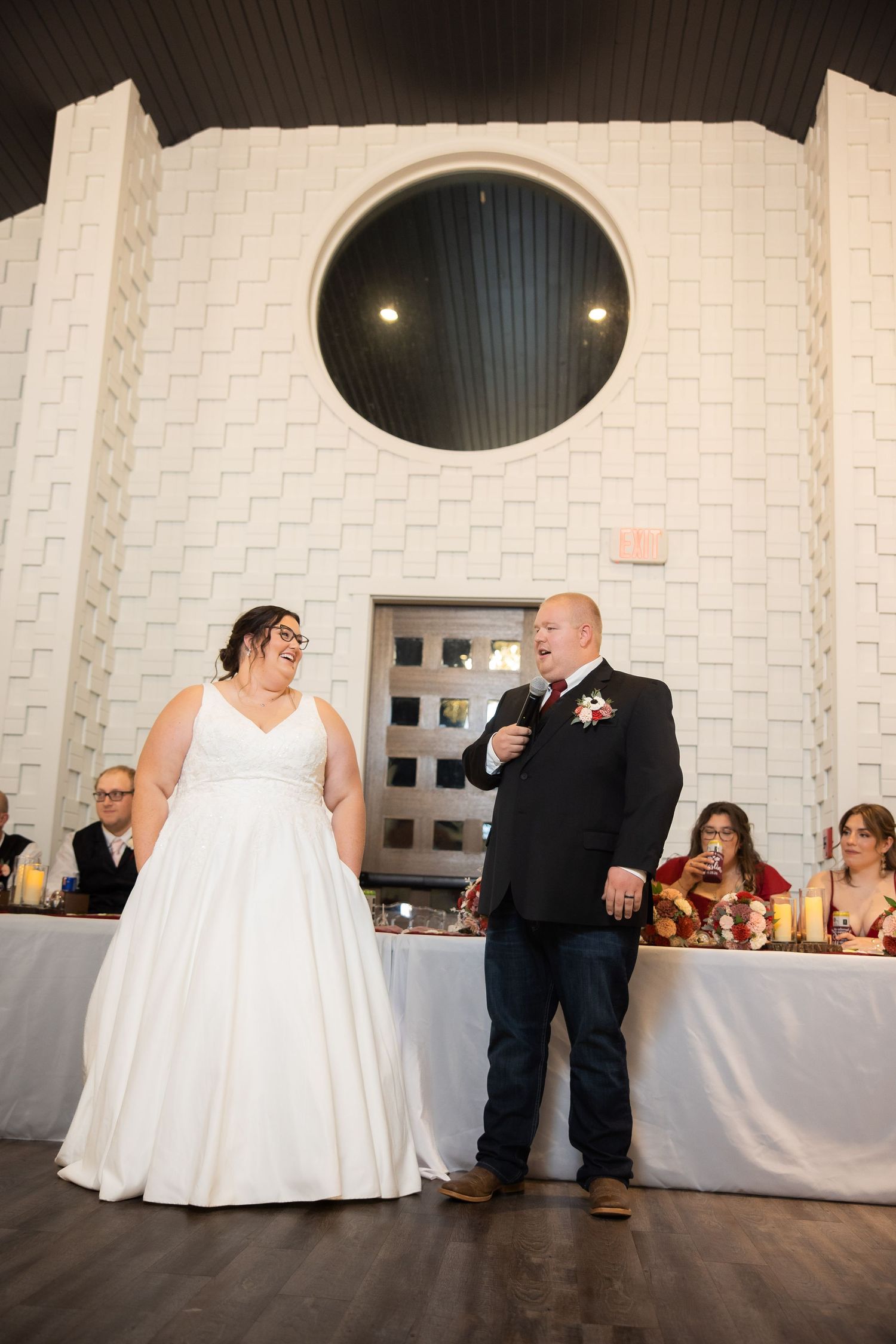 Wedding couple sits together against white textured wall during reception.