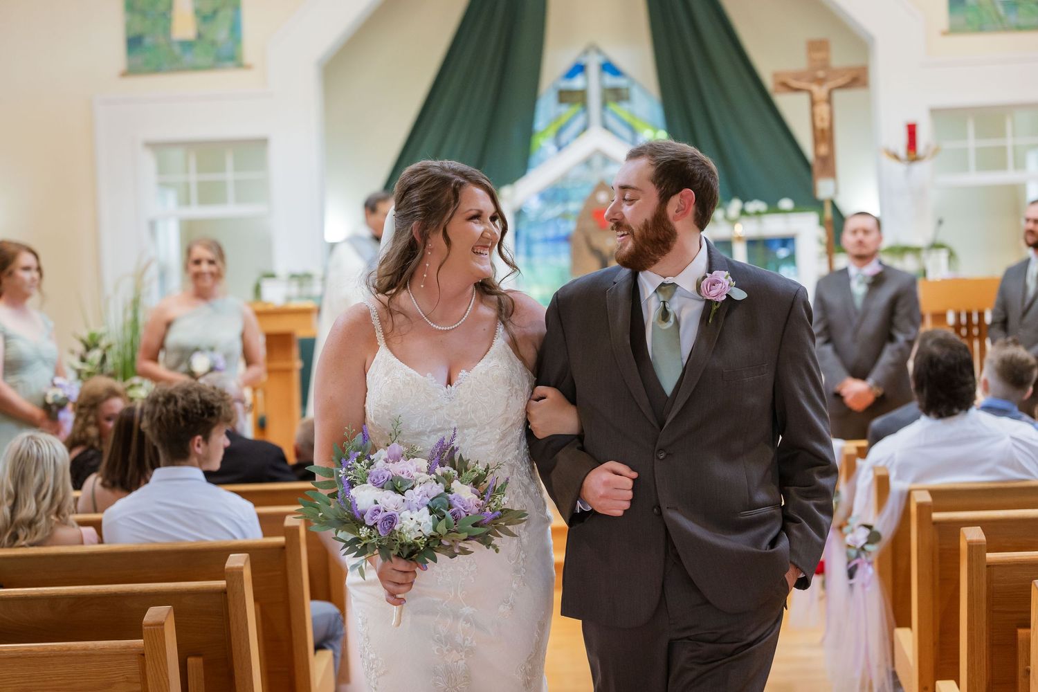 Bride and groom walk down church aisle smiling, bride holding purple bouquet, guests seated in wooden pews.