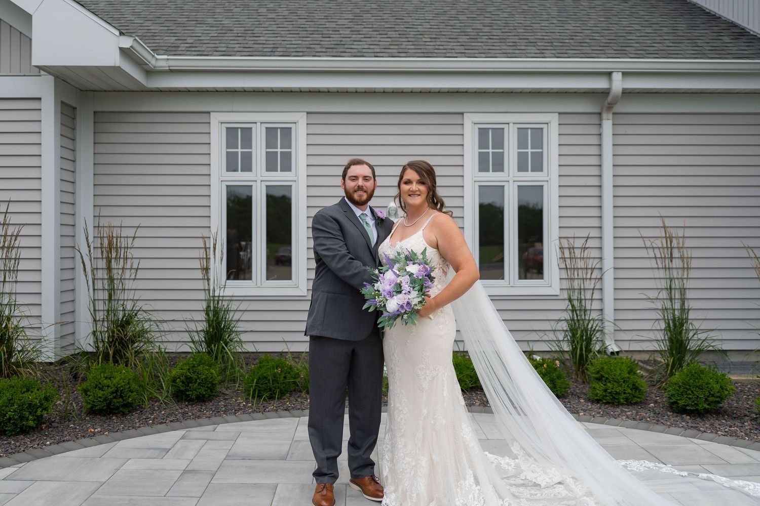 Smiling bride and groom pose together outside gray building, bride in lace gown with flowing veil and purple bouquet.
