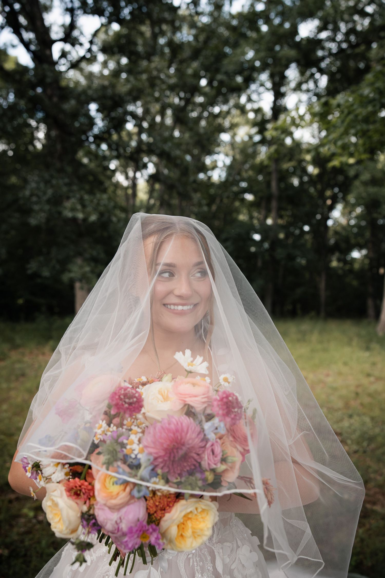Bride wearing veil over face holding vibrant mixed flower bouquet outdoors near lush green trees.