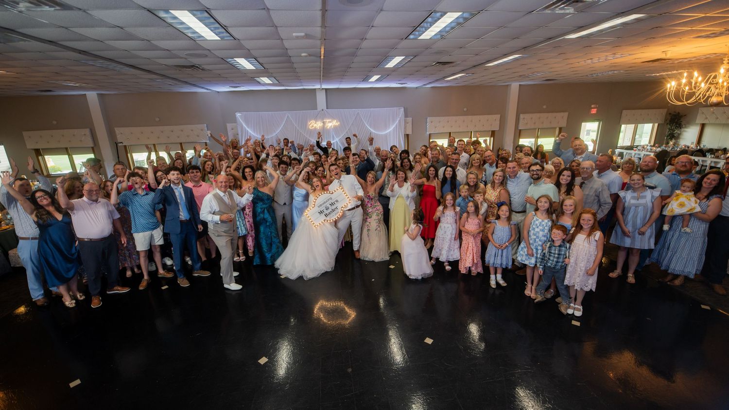 Large wedding group photo with bride and groom kissing at center surrounded by all guests on a dance floor.