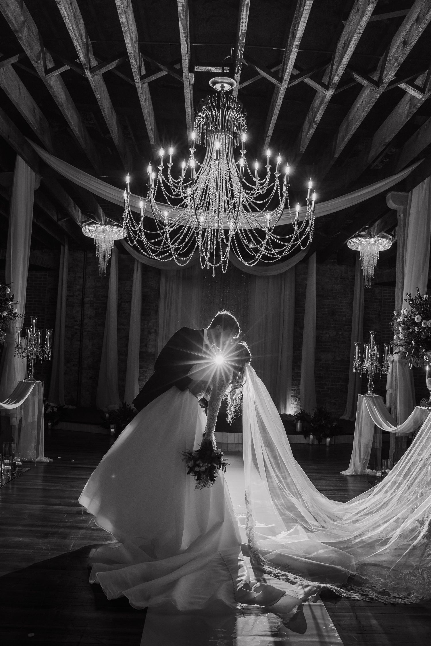 Black and white wedding photo of bride and groom kissing under elegant chandelier in decorated venue.