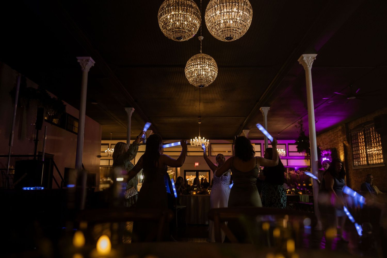 Elegant crystal chandeliers illuminate a dark reception venue with purple accent lighting.