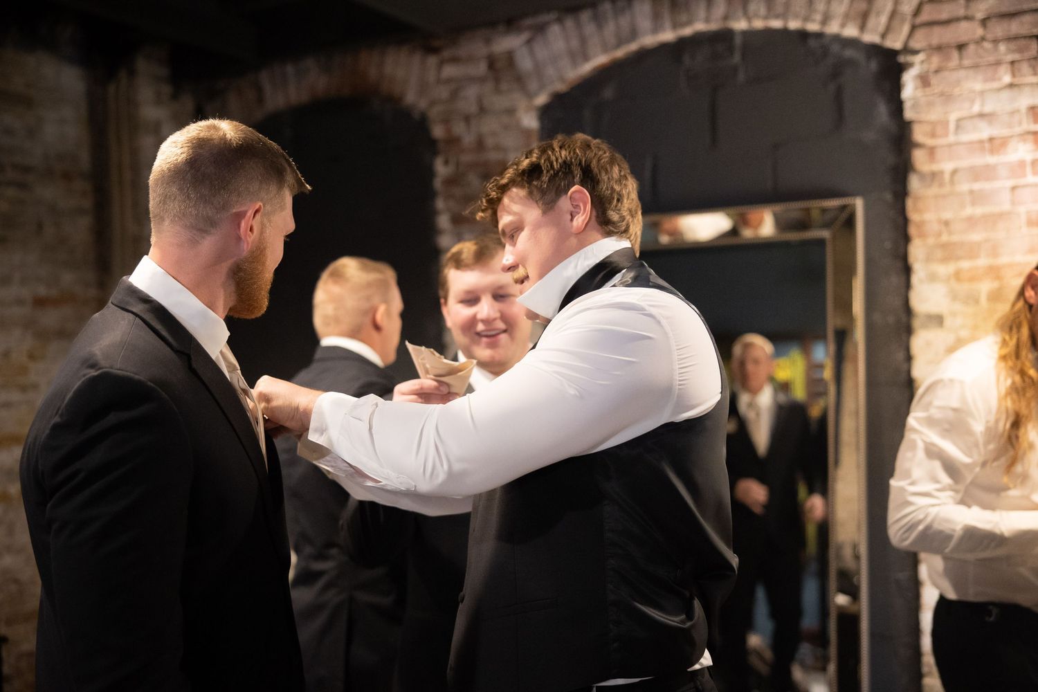 Groomsmen help each other get ready in a rustic brick-walled venue before the wedding ceremony.