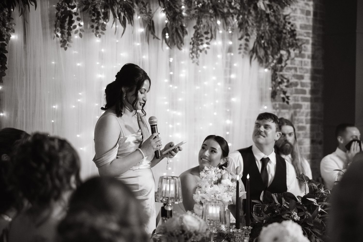 Black and white photo of wedding reception speech with fairy lights and greenery decorations.