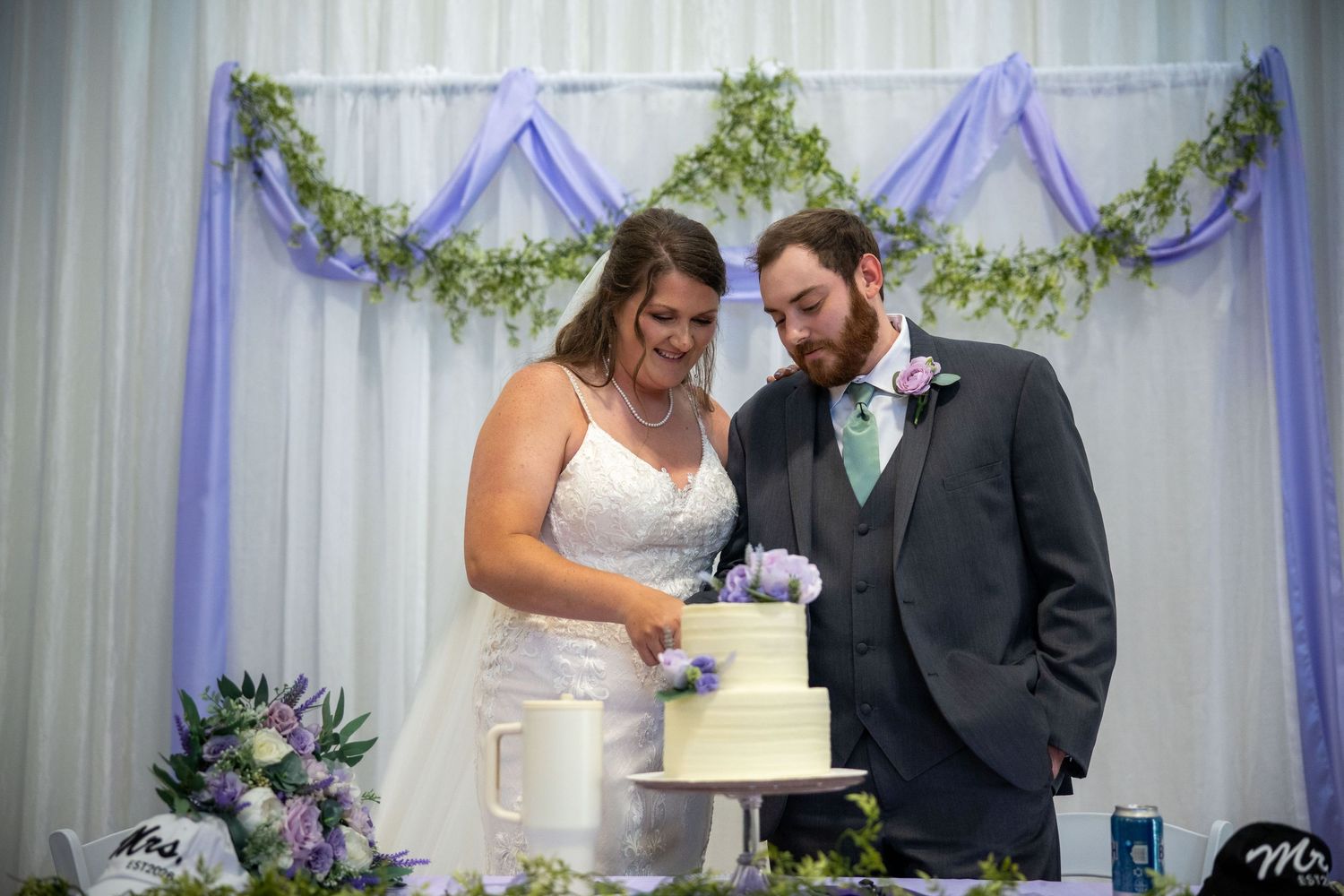 Bride and groom cutting their white wedding cake at a decorated reception venue with purple floral arrangements.