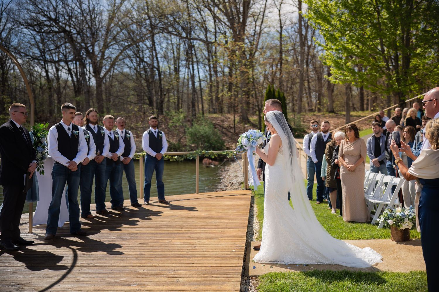 Bride and groom share first kiss on wooden dock aisle surrounded by groomsmen and wedding guests outdoors.