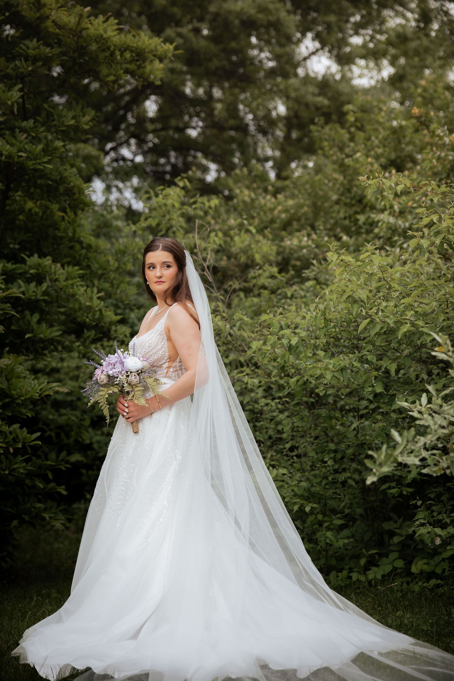 Elegant bride in white ball gown and veil holds purple bouquet in lush green garden setting.