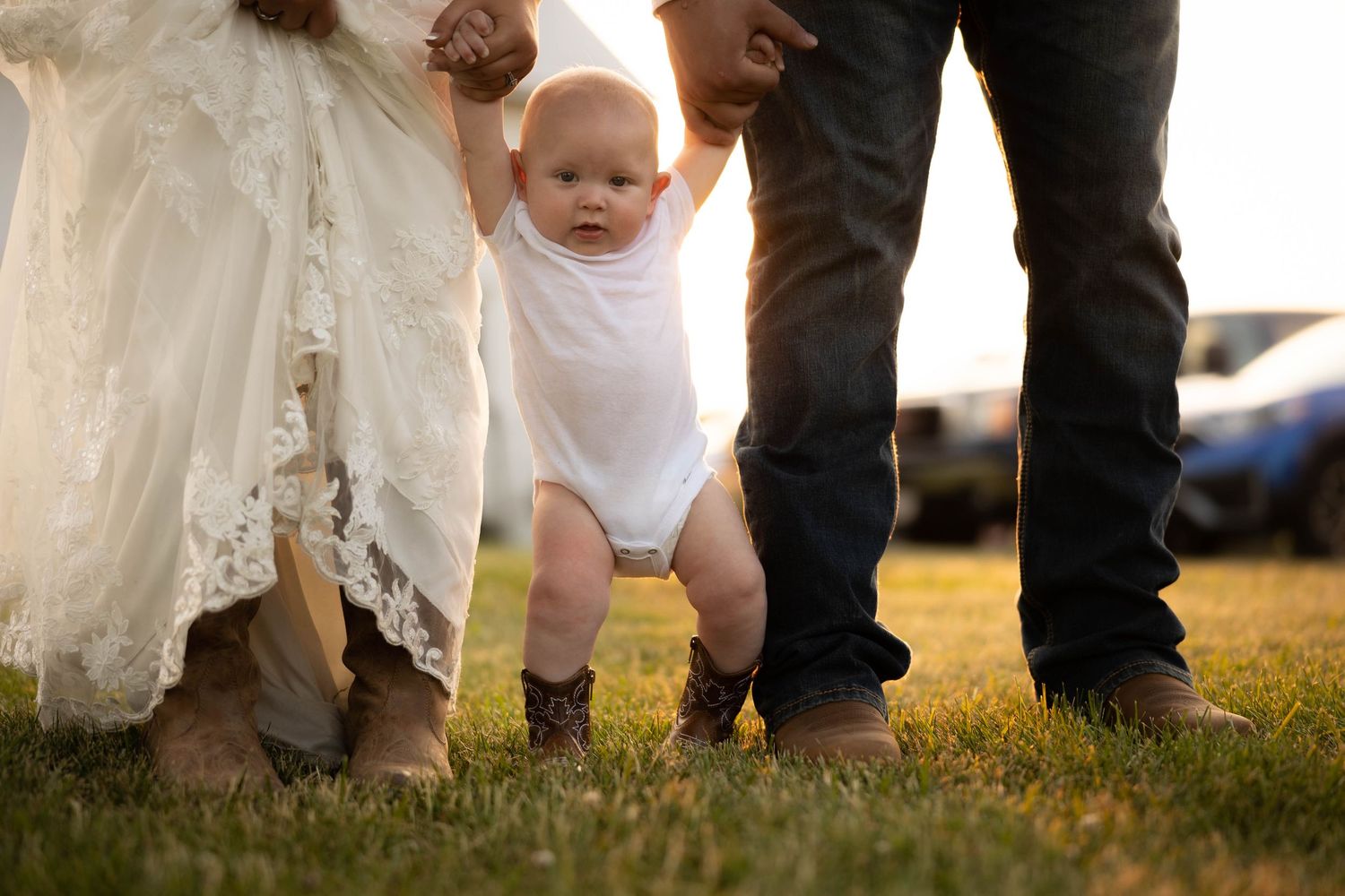 Baby in white onesie and boots takes first steps on grass, held by parents in wedding attire at golden hour.