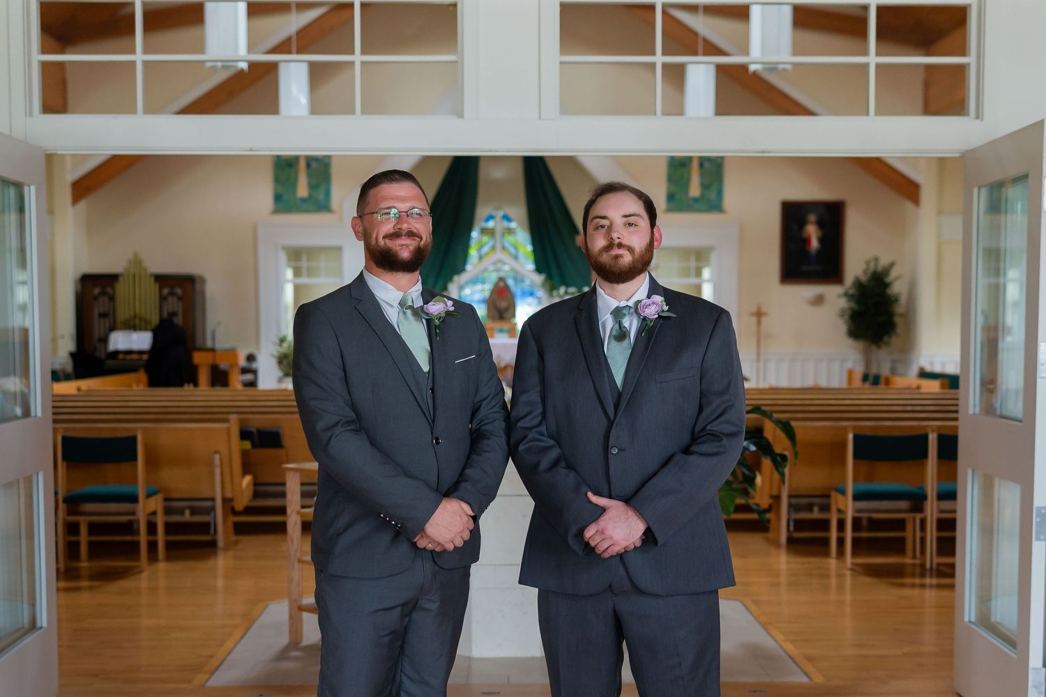 Two groomsmen in gray suits with sage green boutonnieres stand inside a white church with wooden pews.