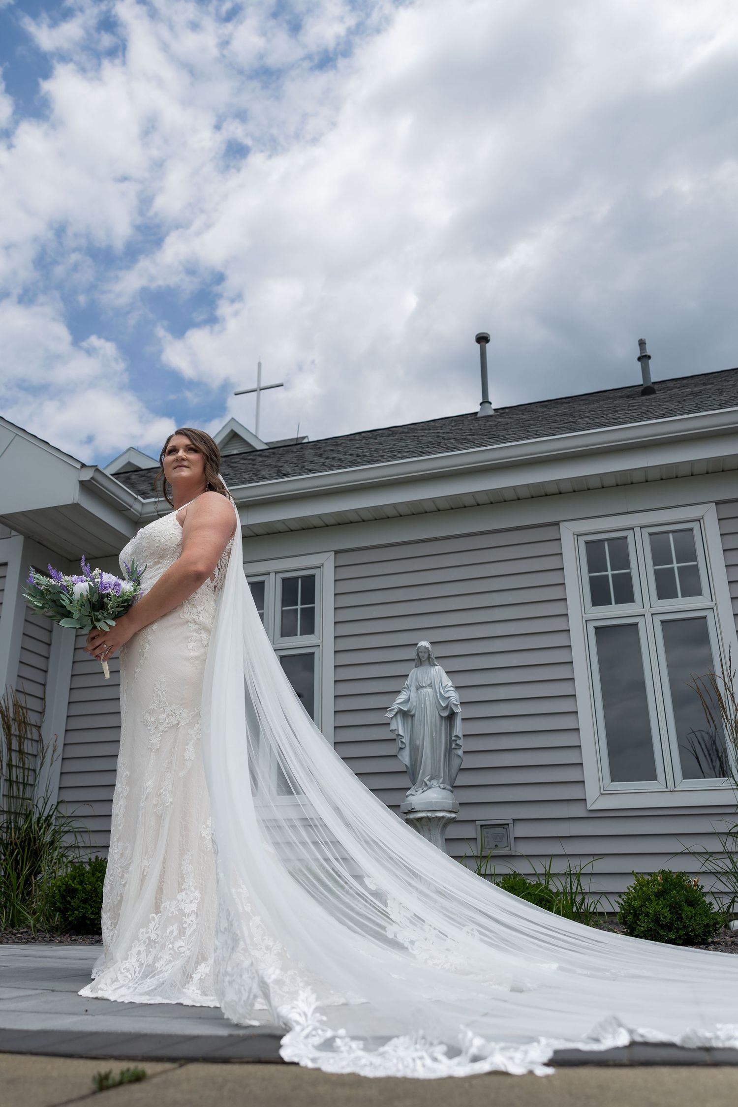 Elegant bride in lace gown with cathedral veil posing before church with Virgin Mary statue.
