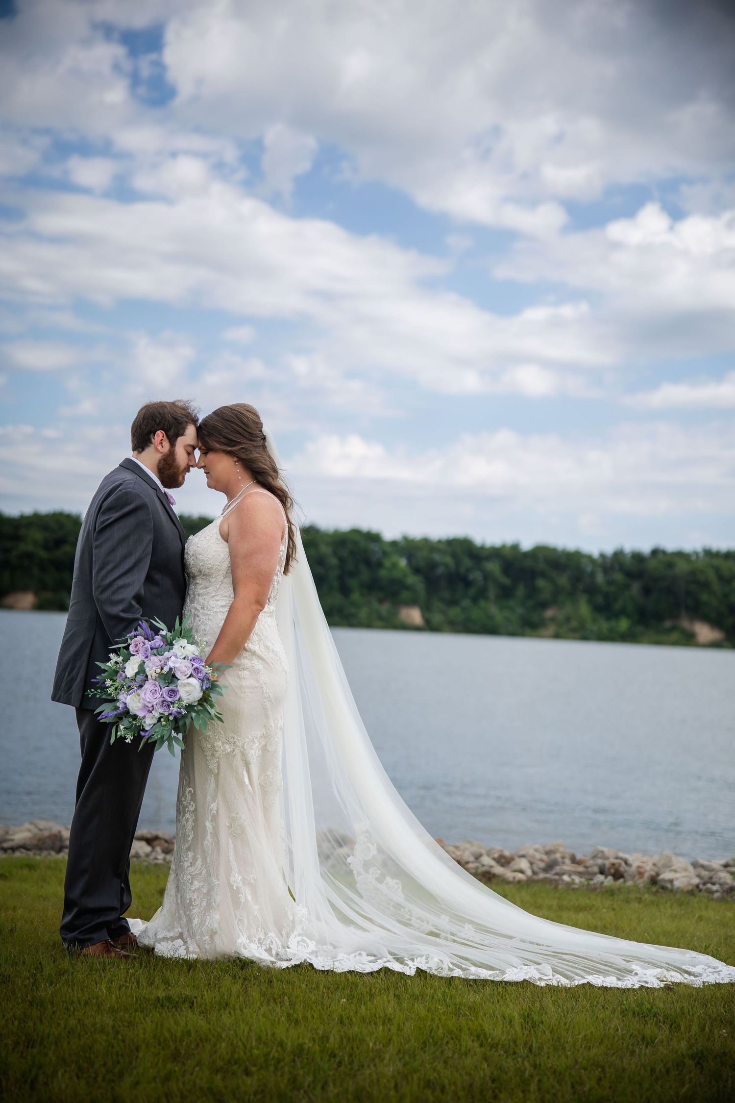 Bride and groom share an intimate moment by a lakeside, her lace gown and veil trailing on green grass.