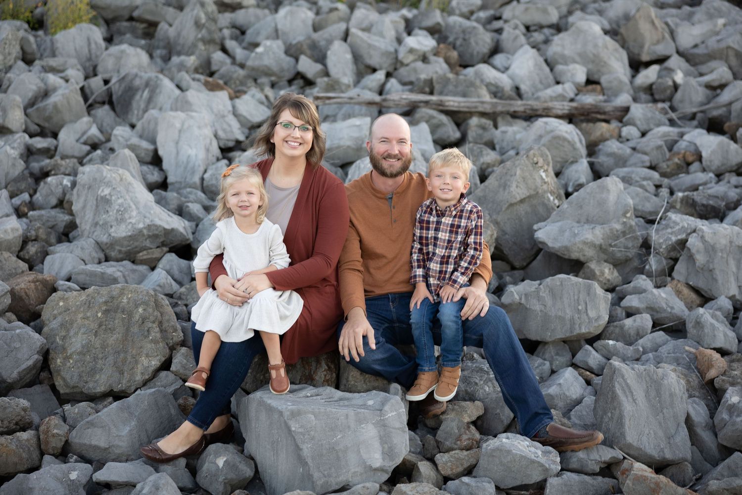 A happy family of four sits together on rocky terrain for a casual outdoor portrait.