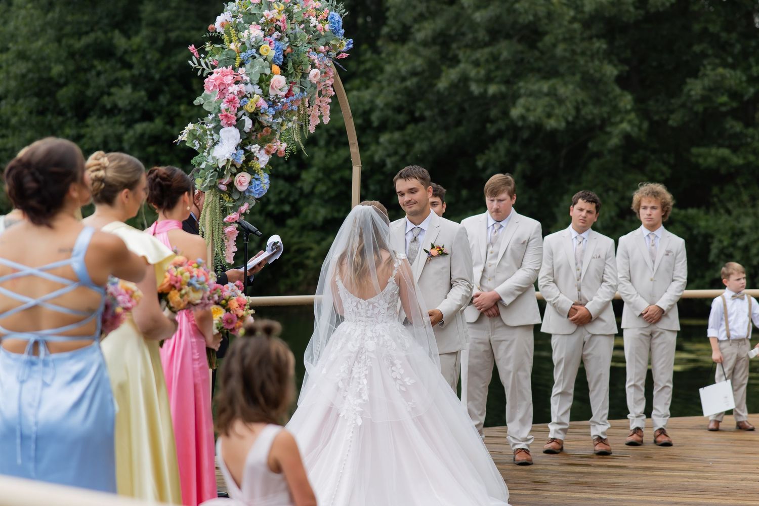 Bride walks down the aisle toward groom and groomsmen at outdoor wedding ceremony with floral arch.