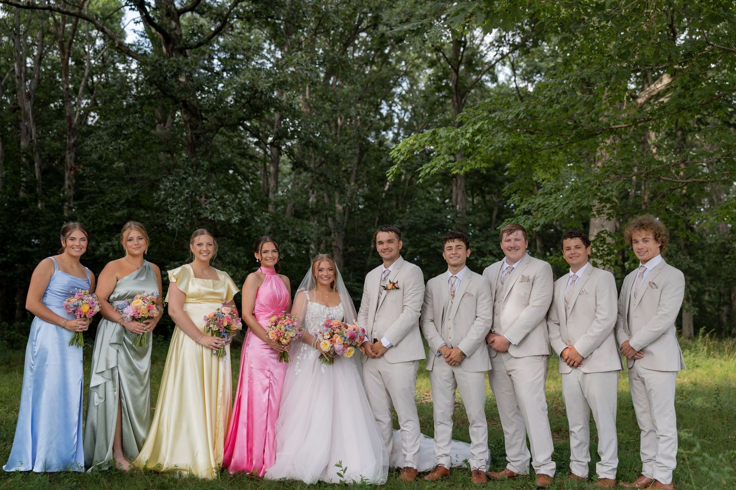 Wedding party posing outdoors, bridesmaids in colorful gowns and groomsmen in tan suits, surrounded by lush green trees.