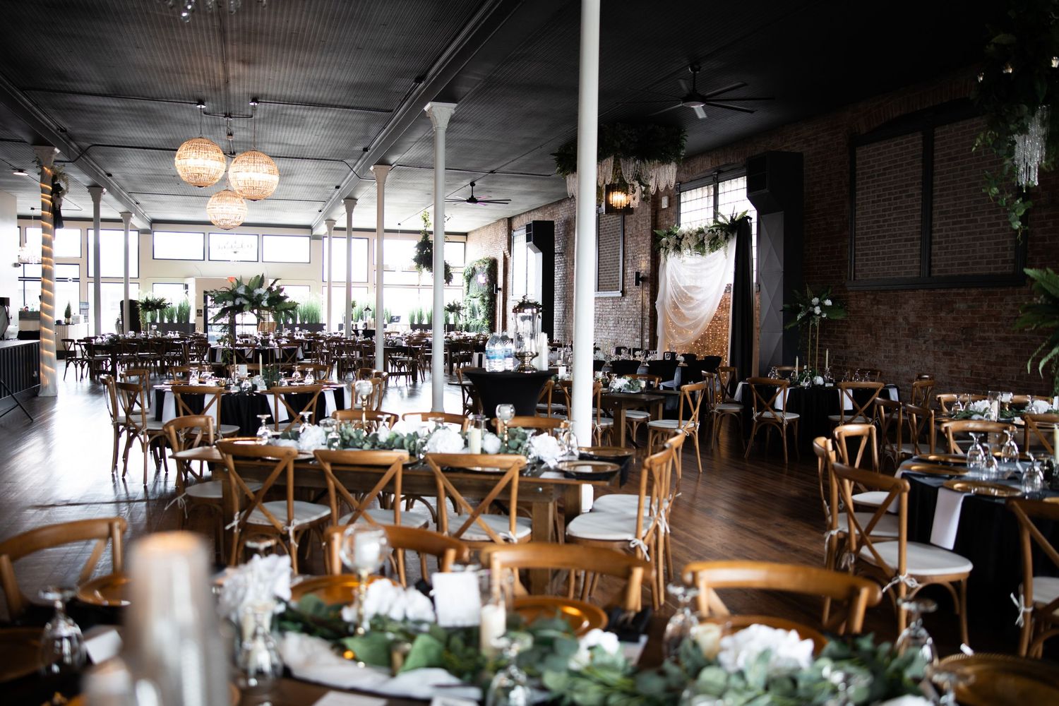 Elegant wedding reception venue with round tables, wooden chairs, greenery centerpieces, and industrial brick walls with pendant lights.