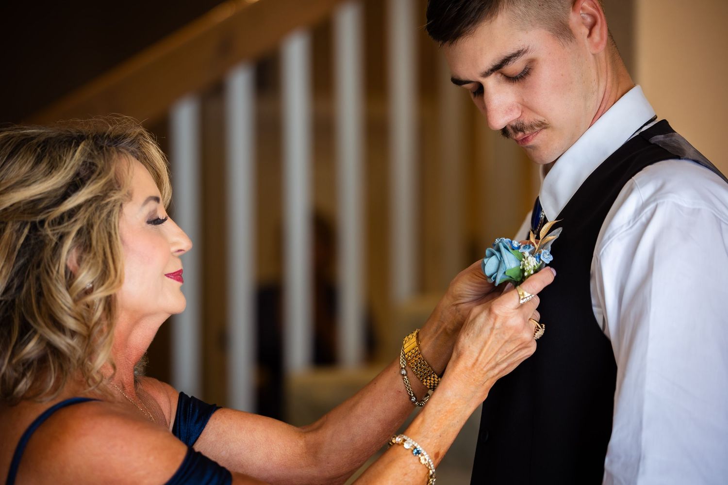 A woman pins a blue boutonniere on a young man in a black vest and tie at a formal wedding event.