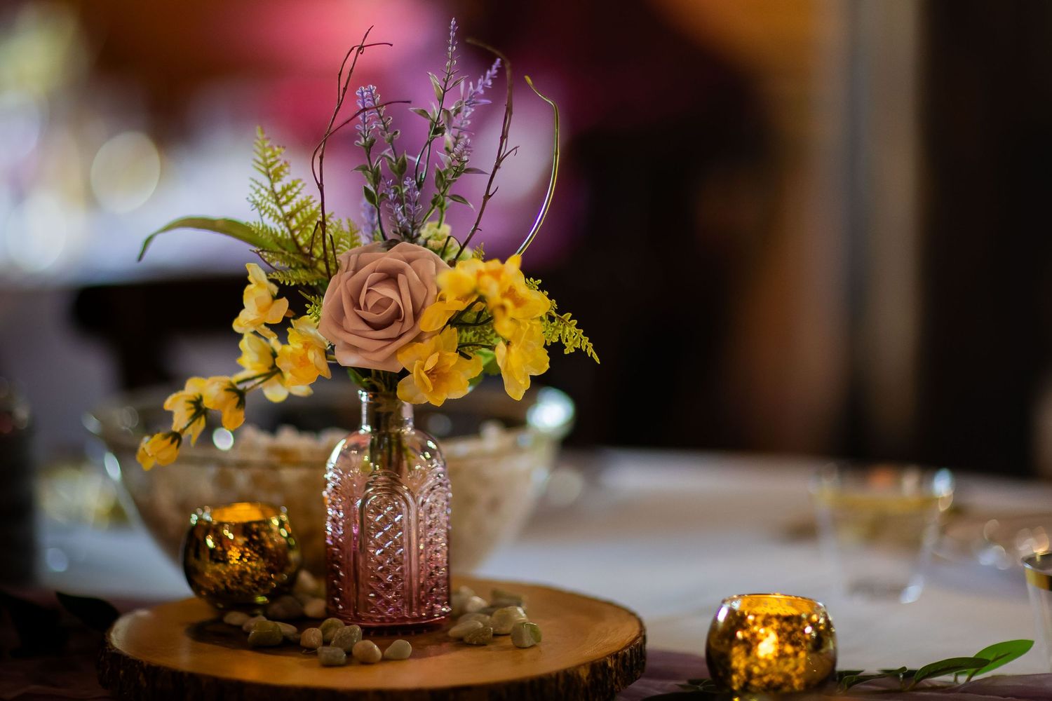 Rustic wedding centerpiece featuring pink and yellow flowers in a vintage glass bottle on a wooden slice.