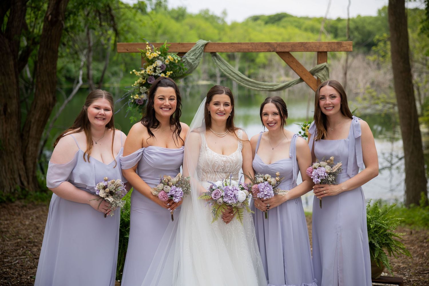 Bride poses with four bridesmaids in matching lavender gowns holding bouquets near a rustic arch by a river.