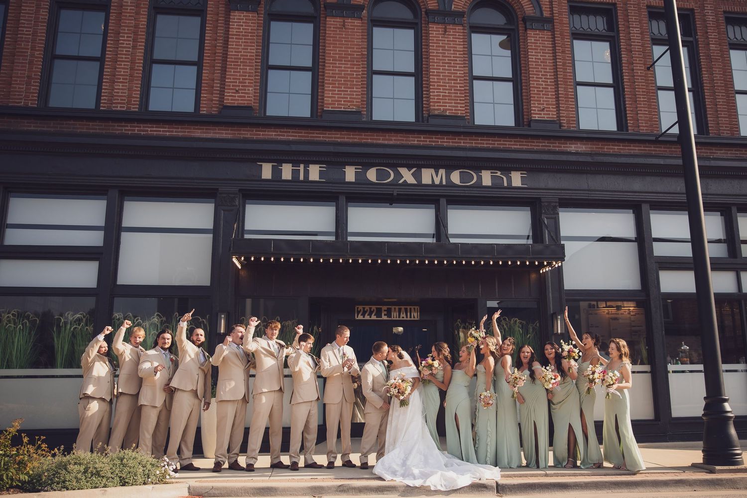 Wedding party celebrating outside The Foxmont venue, bridesmaids in sage green and groomsmen in tan suits cheering with raised arms.