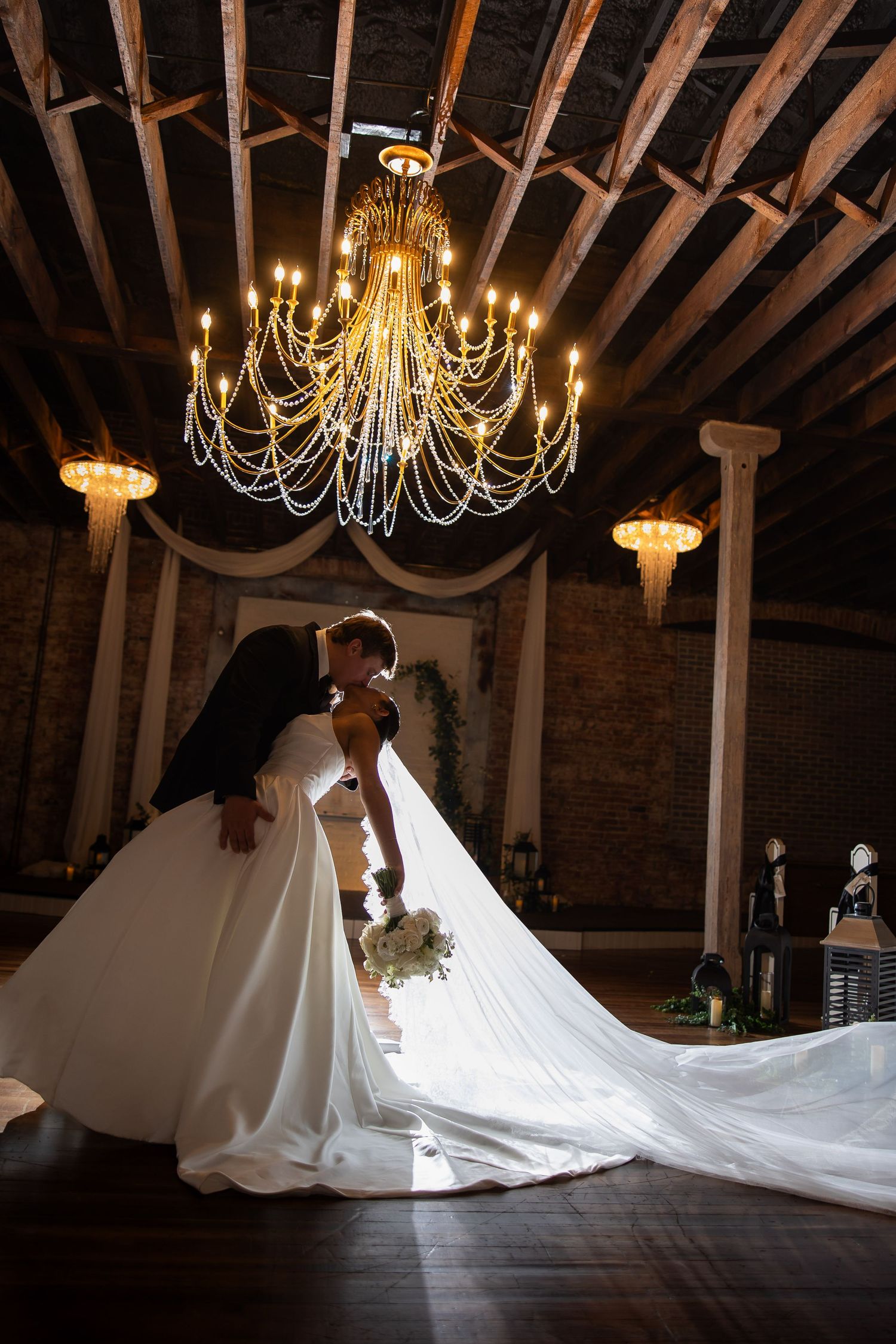 Dramatic wedding photo under vintage chandeliers with a flowing white dress train in an industrial venue.