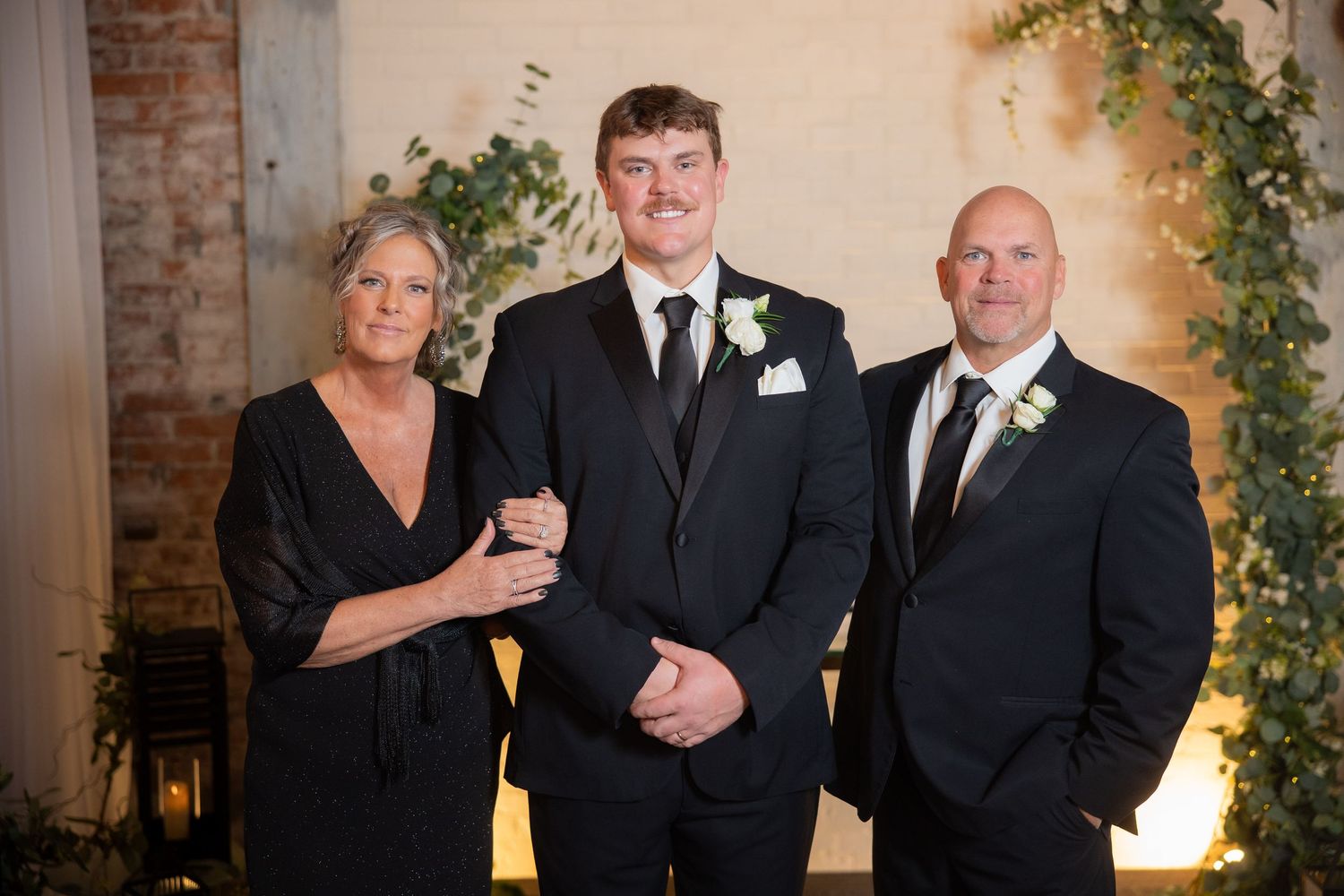 Three people in formal attire pose together at an elegant wedding venue with exposed brick and floral decor.