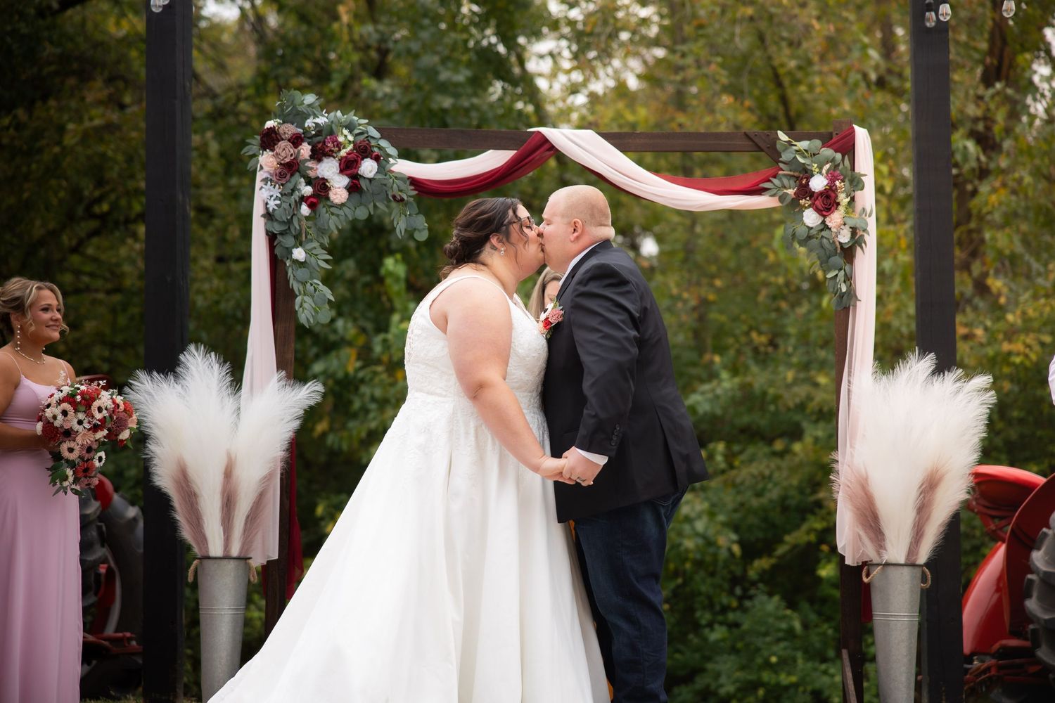 Wedding ceremony takes place in lush outdoor setting with white draped arch and burgundy floral decorations.