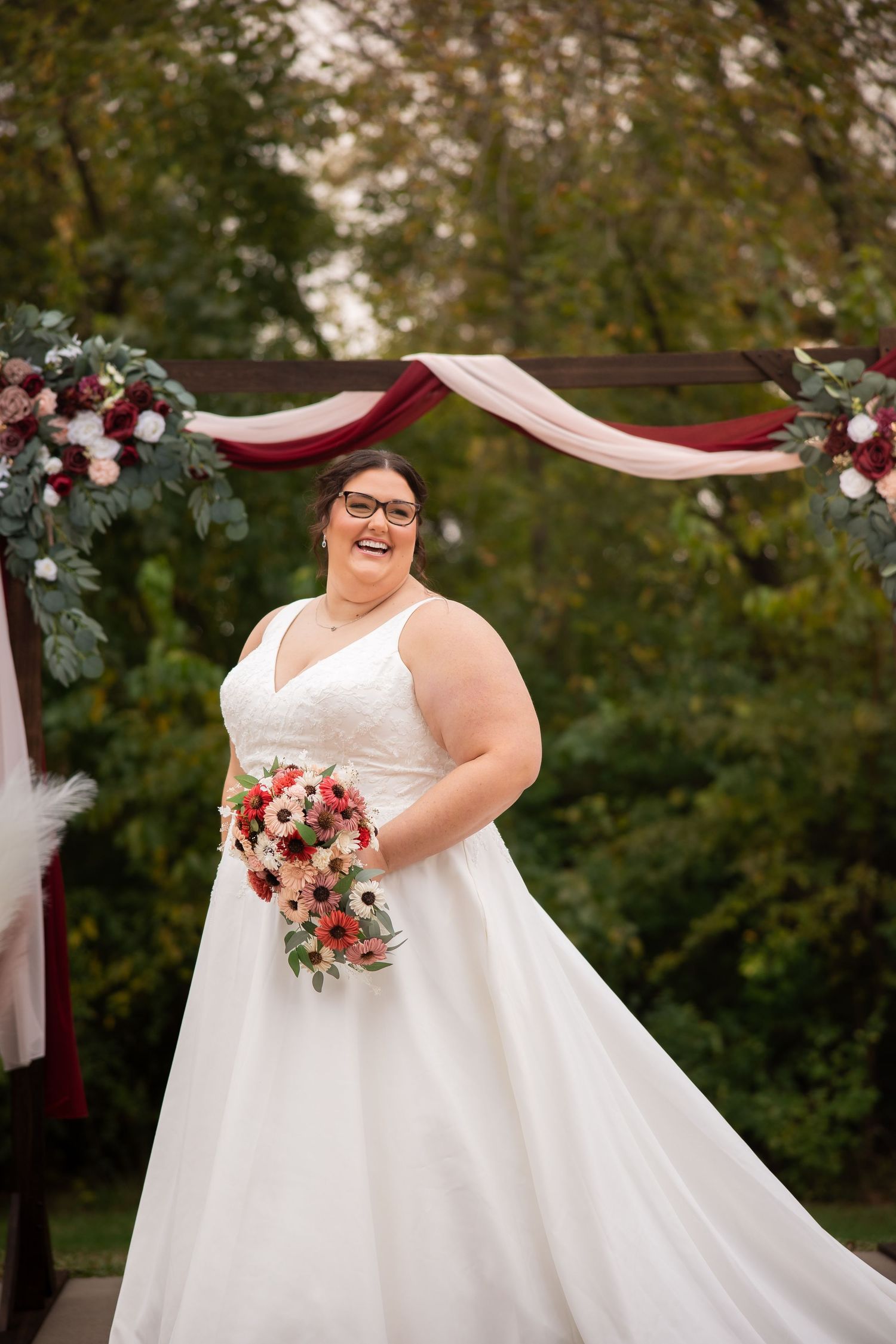 A bride in a white wedding dress holds a burgundy and pink bouquet while posing outdoors under a decorated arch.
