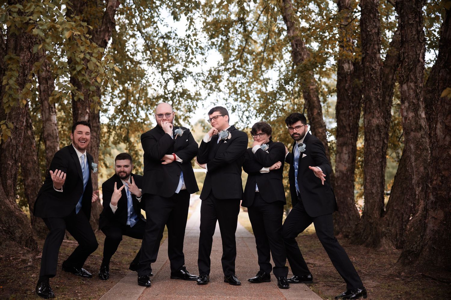 Groomsmen strike playful poses in wooded area during wedding photo session.