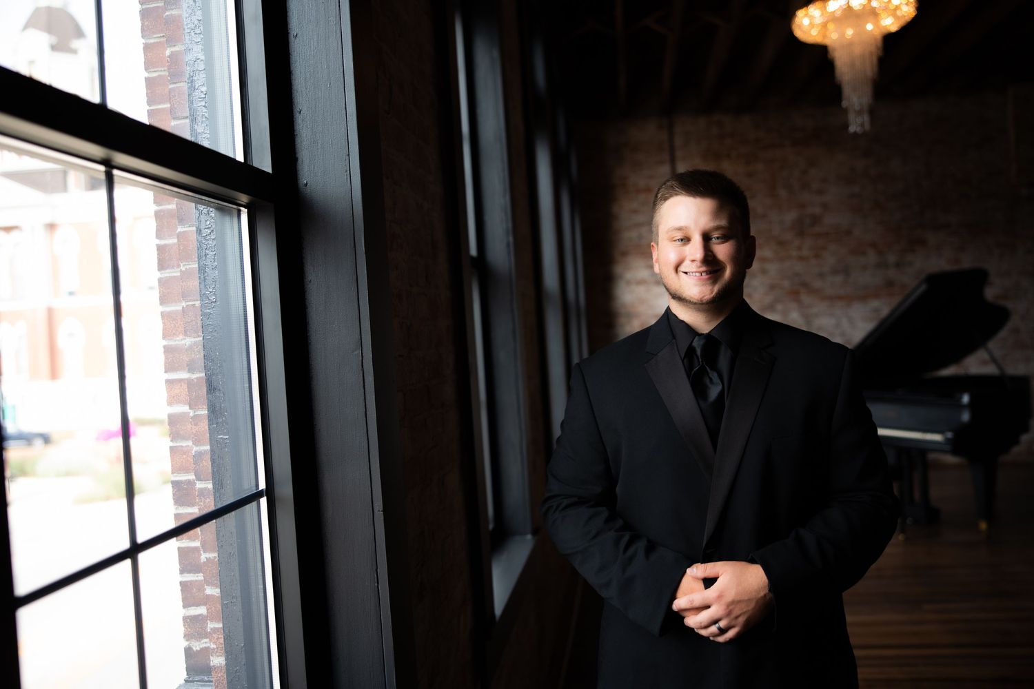Young man in black suit smiling near window with grand piano and chandelier in elegant brick venue.