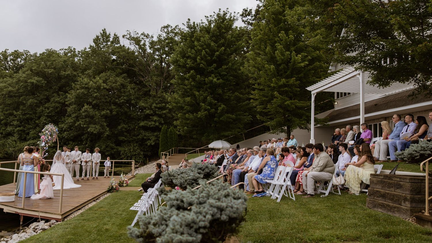 Outdoor wedding ceremony on a lakeside lawn with guests seated in white chairs surrounded by lush green trees.