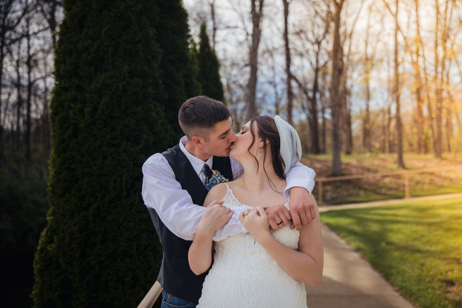Bride and groom sharing a romantic kiss outdoors at golden hour near evergreen trees on their wedding day.