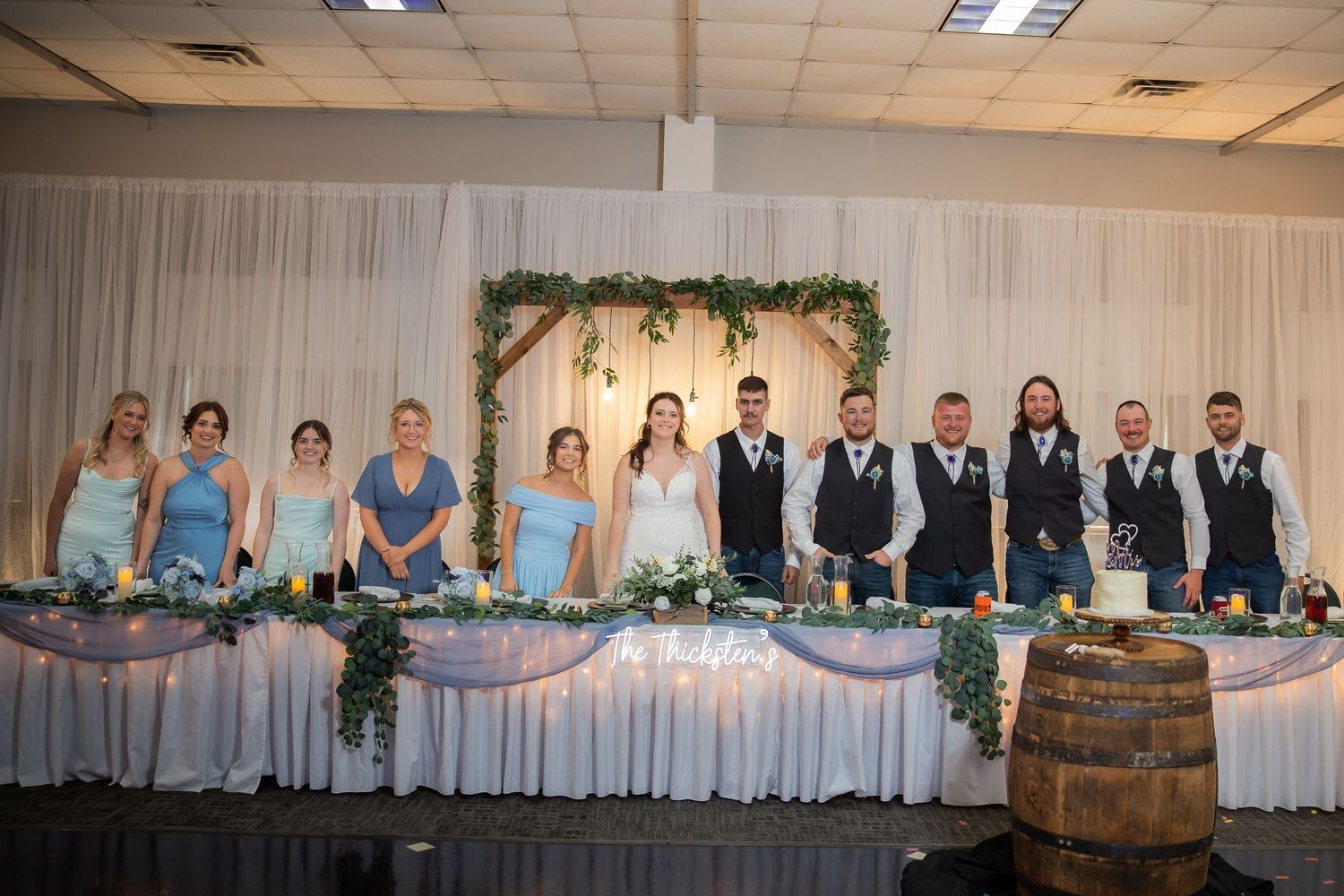 Wedding party poses at decorated head table with greenery arch and barrel accents at reception.